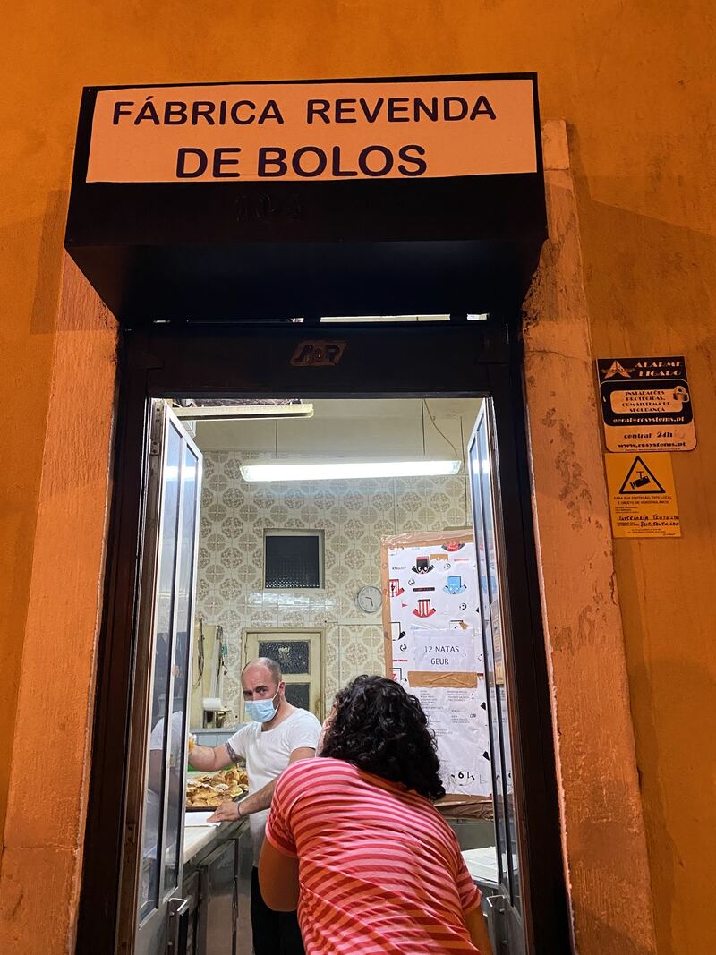 A porta da fábrica aberta à noite, com o letreiro iluminado por cima, um cliente ao balcão e um pasteleiro com um tabuleiro de bolos.